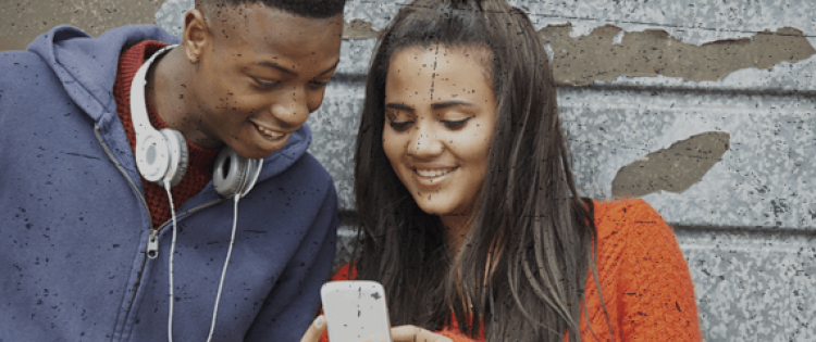 Teenage boy with headphones around neck and teenage girl holding phone. both are looking at the phone and smiling