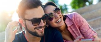 Photo of a man sitting outside on steps listening to headphones. His girlfriend sits next to him, holding her phone.
