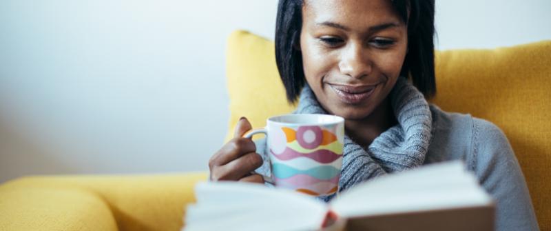 Good Book Photo of a woman reading a book with a mug of tea. She's sitting in a yellow chair.