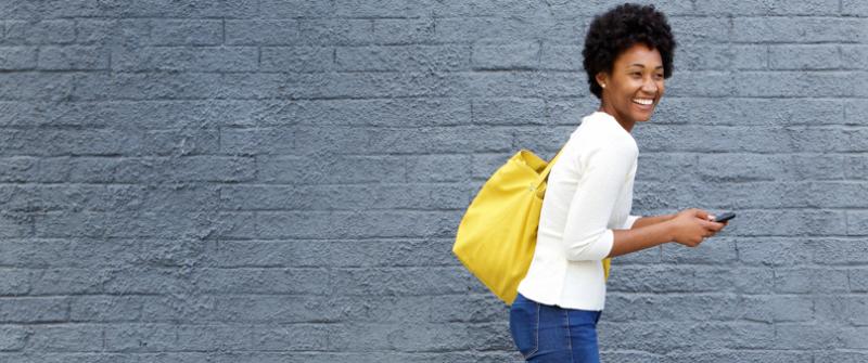 Woman with a Yellow Bag Photo fo a smiling woman carrying a yellow bag. She's walking in front of a gray brick wall and carrying her phone.