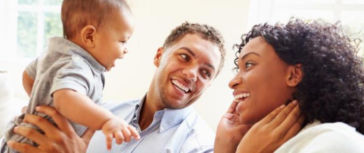 Photo of a smiling mom, dad, and baby boy. Dad is holding the baby up and gazing lovingly at mom.