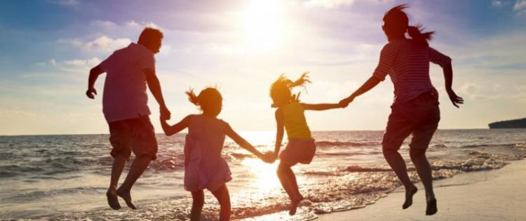Photo of mom, dad, and two girls holding hands and jumping into the surf on the beach. The sun is setting behind the family.