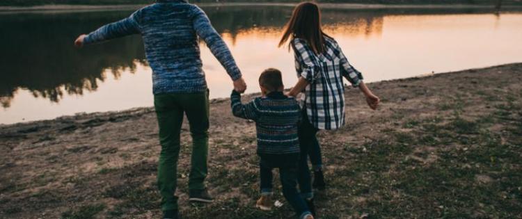 Photo of two adults with a child, walking towards a river and holding hands.