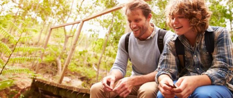 Photo of a dad chatting with his son. They are sitting on a wooden bridge along a forest trail.