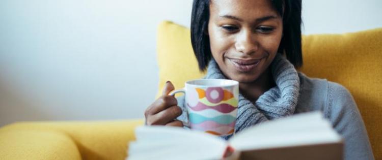 Photo of a woman reading a book with a mug of tea. She is sitting in a yellow chair.