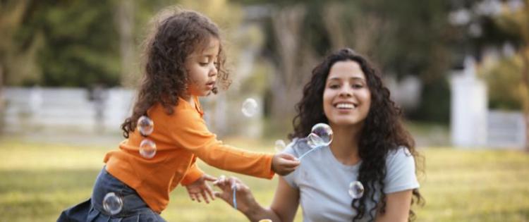 Photo of a mom helping her daughter blow bubbles in the park.