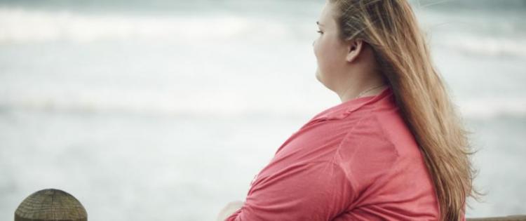 Photo of a young woman with her back turned. She's standing at a fence, gazing out at the ocean.