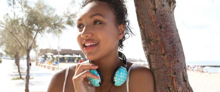 Photo of a smiling young woman leaning against a tree. She has polka dot headphones around her neck.