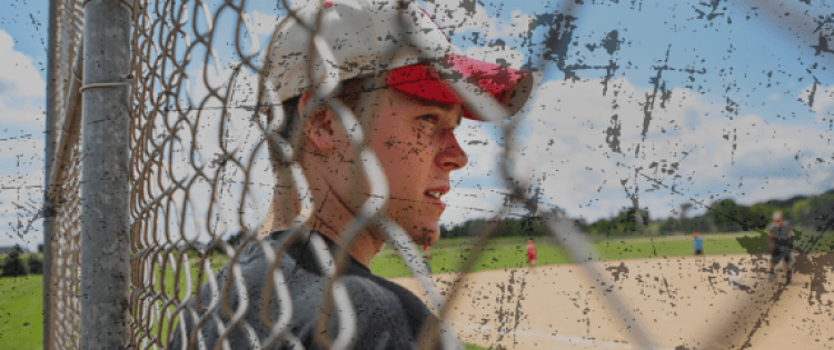 Teenage boy in baseball cap standing in front of fence facing a baseball field