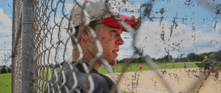 Teenage boy in baseball cap standing in front of fence facing a baseball field