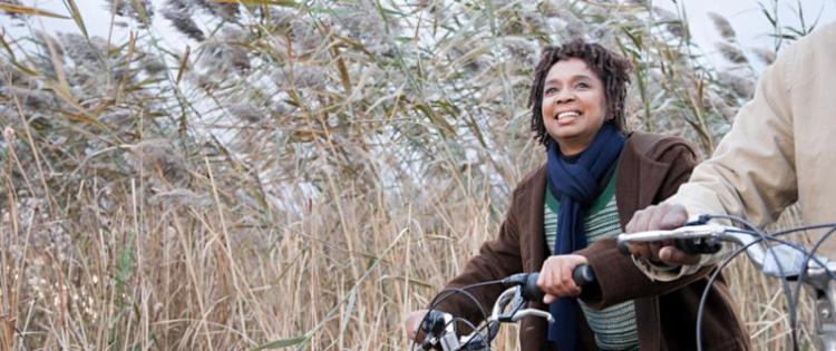 Photo of a woman on a bicycle in autumn, she wears a coat and scarf. A second cyclist can be partially seen to her side.