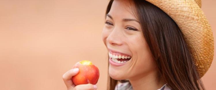Photo of a woman wearing a cowboy-style straw hat. She's smiling and holding a peach that she's take a bite from.