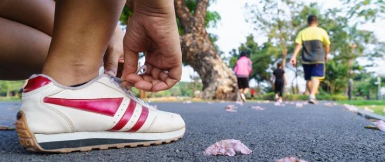 Close-up photo of a person reaching down to lace up a running shoe, other people can be seen walking in the background.