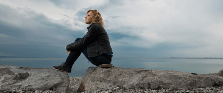 Photo of a blonde woman sitting on a rocky beach on a cloudy day.