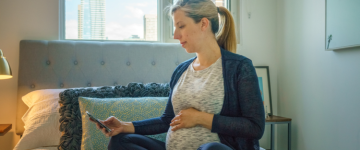 Photo of a blond pregnant woman looking at her smartphone while sitting on her bed