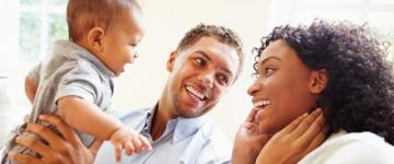 Photo of a smiling mom, dad, and baby boy. Dad is holding the baby up and gazing lovingly at mom.