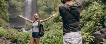 Photo of a man taking a picture of his blonde girlfriend near a waterfall in the jungle