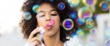 Photo of a woman holding a pink bottle of bubble solution while blowing into a bubble wand. Bubbles are floating off into the air.