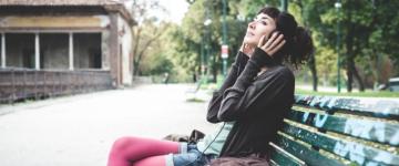 Photo of a woman sitting on a city park bench wearing headphones. Her hands are by her ears, gently pressing the headphones tighter.