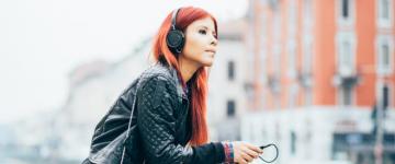 Photo of a young woman with red hair wearing a leather jacket. She's leaning against a stone railing in a city.