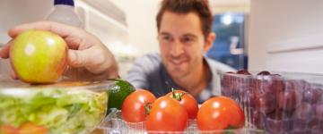 Photo of a man reaching into a fridge full of healthy fruits and vegetables, grabbing an apple.