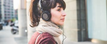 Photo of a woman walking down a city street with headphones and a scarf looks back over her shoulder