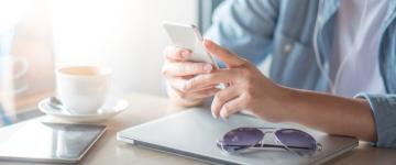 Hands of man using cell phone on coffee table, with laptop, tablet, sunglasses, and coffee on table