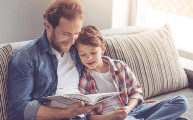 Photo of a man and his young son reading a book together on a sofa