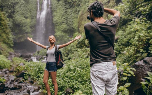 Photo of a man taking a picture of his blonde girlfriend near a waterfall in the jungle