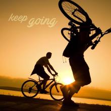 Photo of two men. One is on a bike and one is carrying his bike. Text reads: keep going