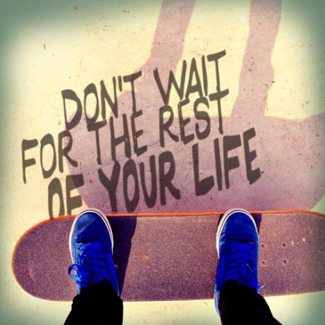 Skate deck/ skate board with feet and text on ground. "Don't wait for the rest of your life."